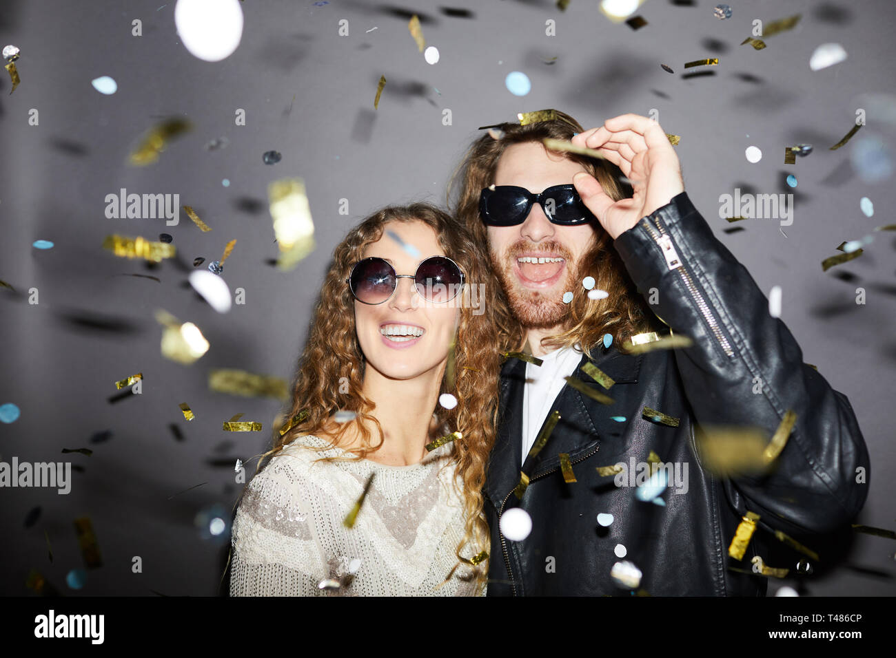 Jeune couple d'hipster joyeux et excité avec des cheveux bouclés portant des lunettes de soleil se tenant sous des confettis dorés et s'amuser à la fête Banque D'Images