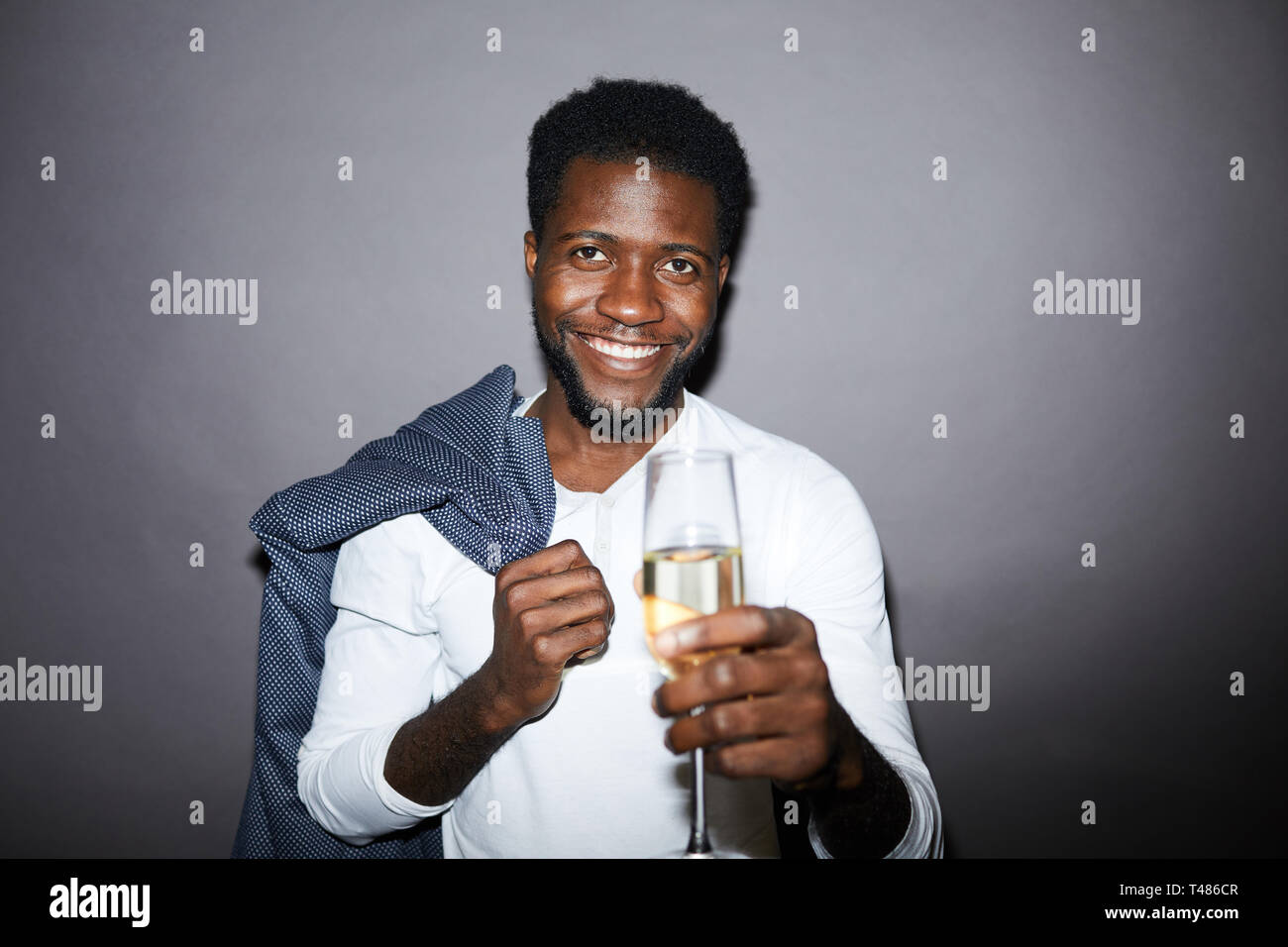 Jeune homme noir souriant avec une veste de maintien pour barbe sur l'épaule et regardant l'appareil photo tout en levant la flûte à champagne Banque D'Images