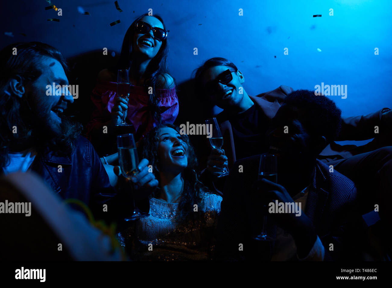 Groupe de jeunes amis positifs riant ensemble et buvant du champagne dans la chambre sombre avec lumière bleue, fille excitée de tomber confetti Banque D'Images