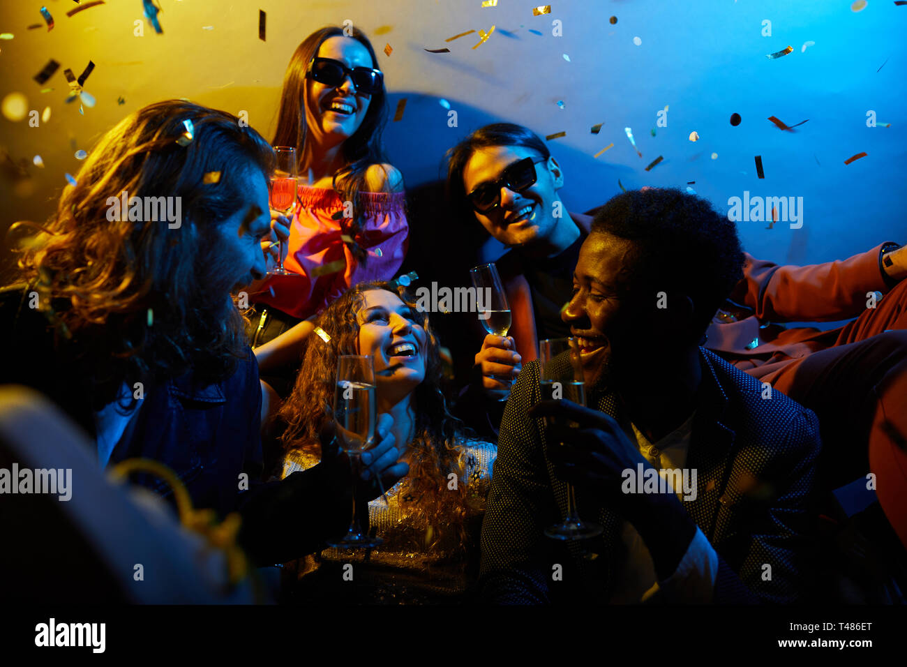 Groupe de jeunes amis multiethniques excités assis ensemble sous confetti et boire de l'alcool tout en riant et en parlant à la fête Banque D'Images