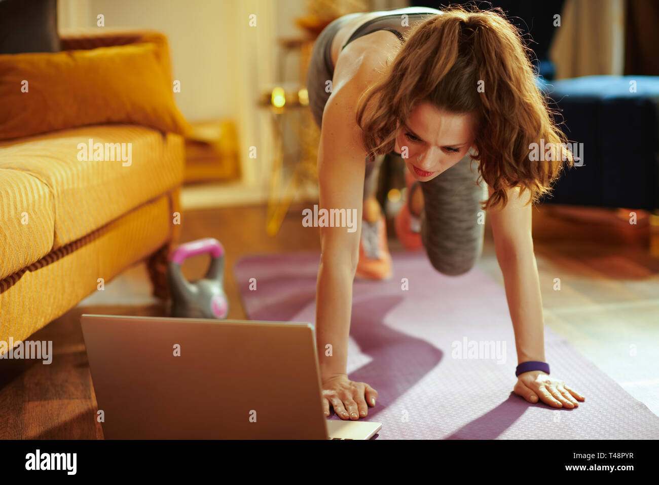 Jeune femme en vêtements de sport fitness à domicile à l'aide de modernes streaming en ligne site de remise en forme et d'ordinateur portable en faisant tapis de sol fitness formation sur circuit. Banque D'Images