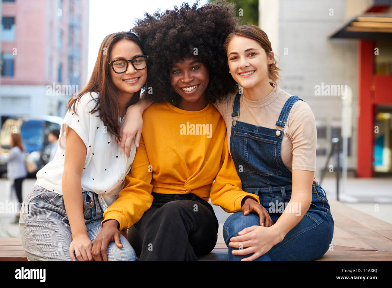 Trois jeunes adultes branchés copines sur un banc dans une rue de ville souriant pour appareil photo, Close up Banque D'Images