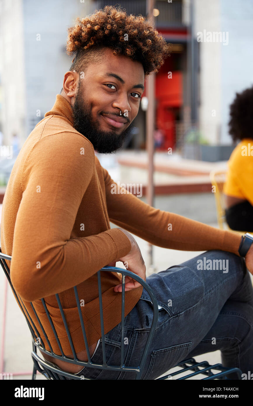 Hipster noir millénaire homme assis à l'extérieur d'un café dans la rue, se retourne en souriant à la caméra, Close up Banque D'Images