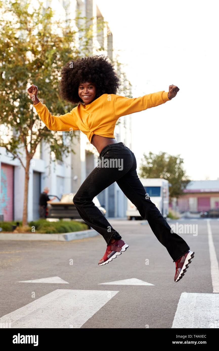 Jeune femme noire afro avec sauts, à la recherche d'appareil photo, pendant la traversée de rue, vue latérale, verticale Banque D'Images
