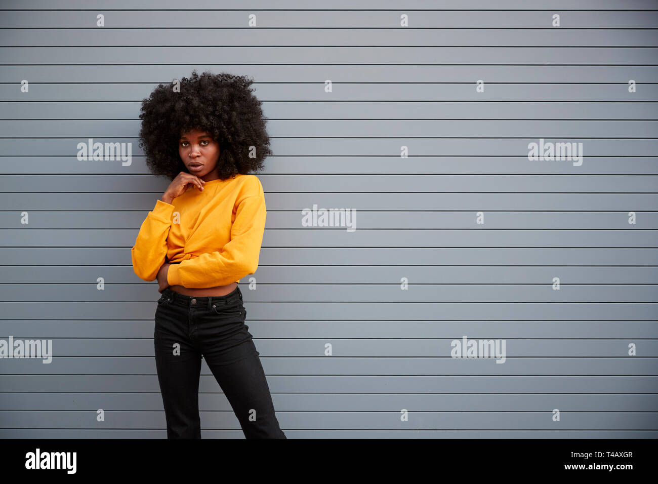 Jeune femme noire de la hanche contre permanent sécurité volets gris, tête sur la main, à la recherche d'appareil photo Banque D'Images