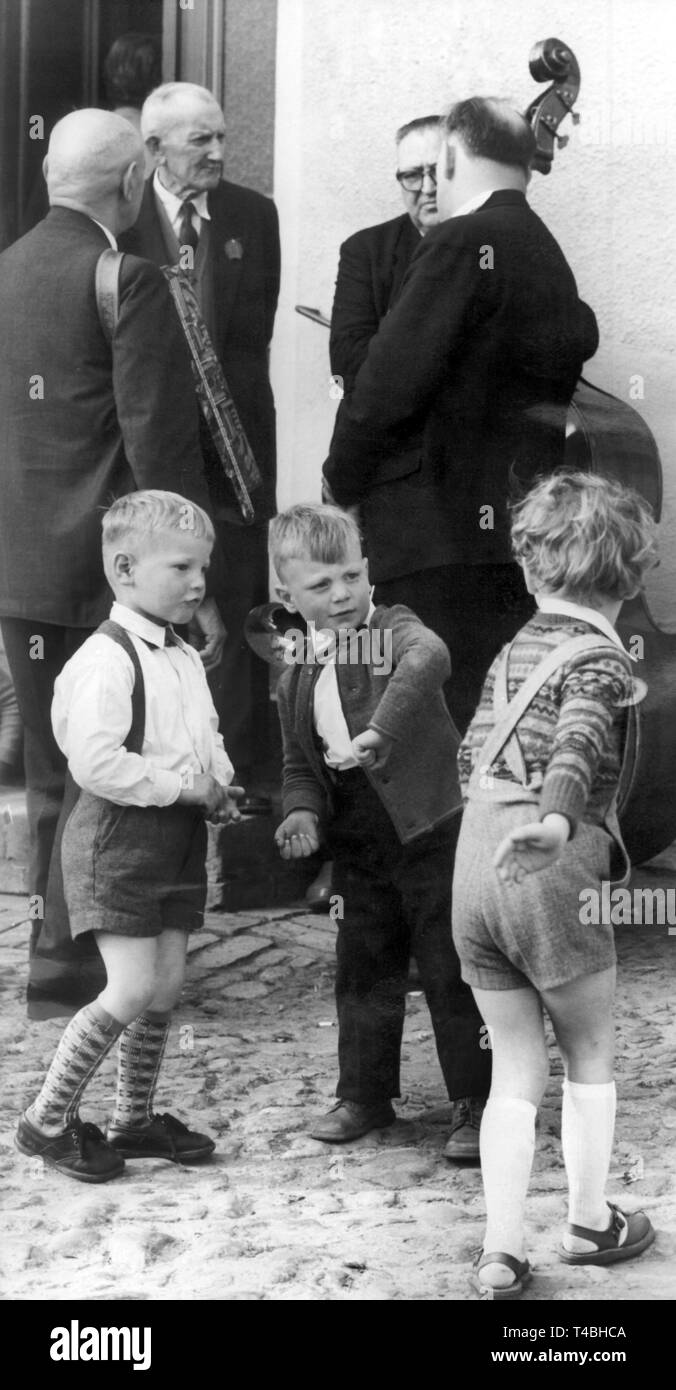 Drei Jungen bei einer Rangelei, während sich im Hintergrund eine Gruppe älterer und Orchester unterhält Mitglieder. Aufnahme vom Mai 1963. Foto : ADN | Le monde d'utilisation Banque D'Images