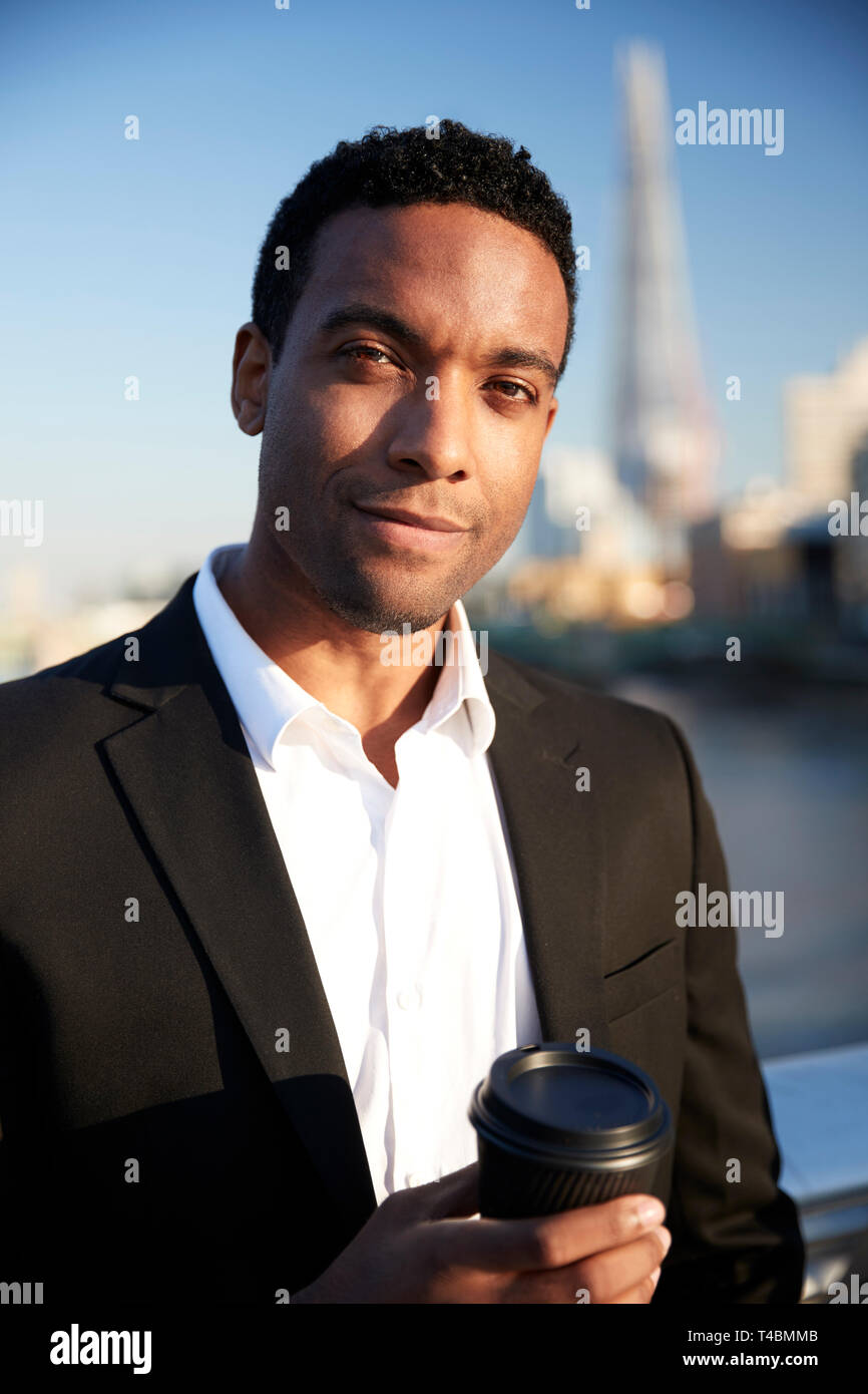 Jeune black businessman dans smart casual clothing debout près de la Tamise à Londres la tenue d'un café à emporter, à à l'appareil photo, Close up Banque D'Images
