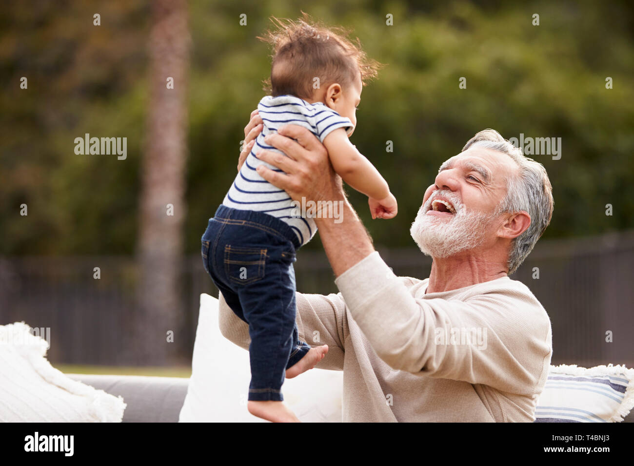 Senior homme assis dans le jardin de lever son petit-fils bébé dans l'air et souriant Banque D'Images