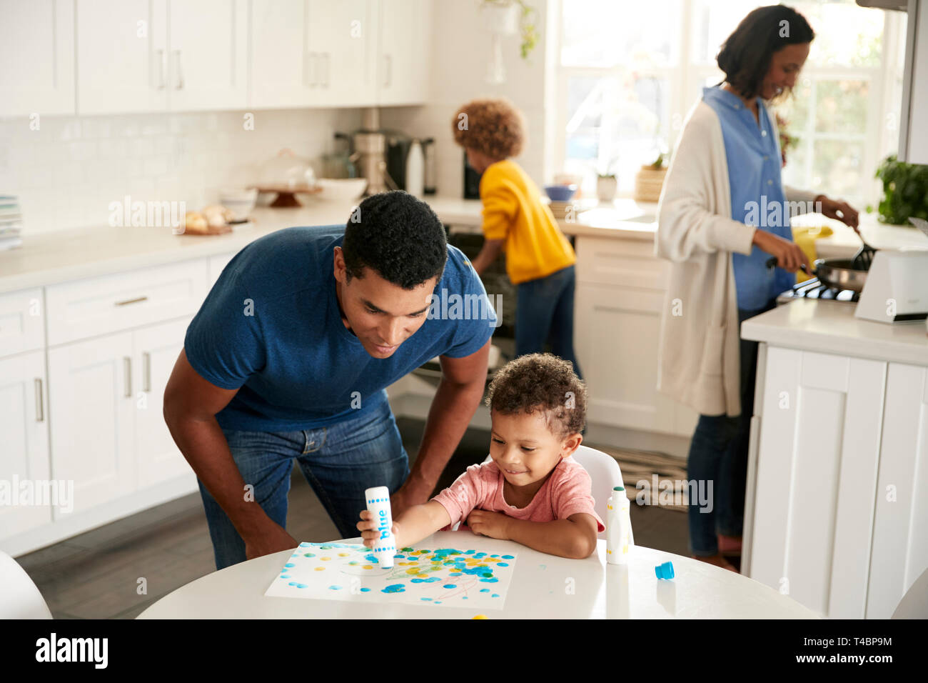 Papa regarder son petit garçon une peinture photo assis à une table dans la cuisine, tandis que mère et fille préparer la nourriture dans l'arrière-plan Banque D'Images