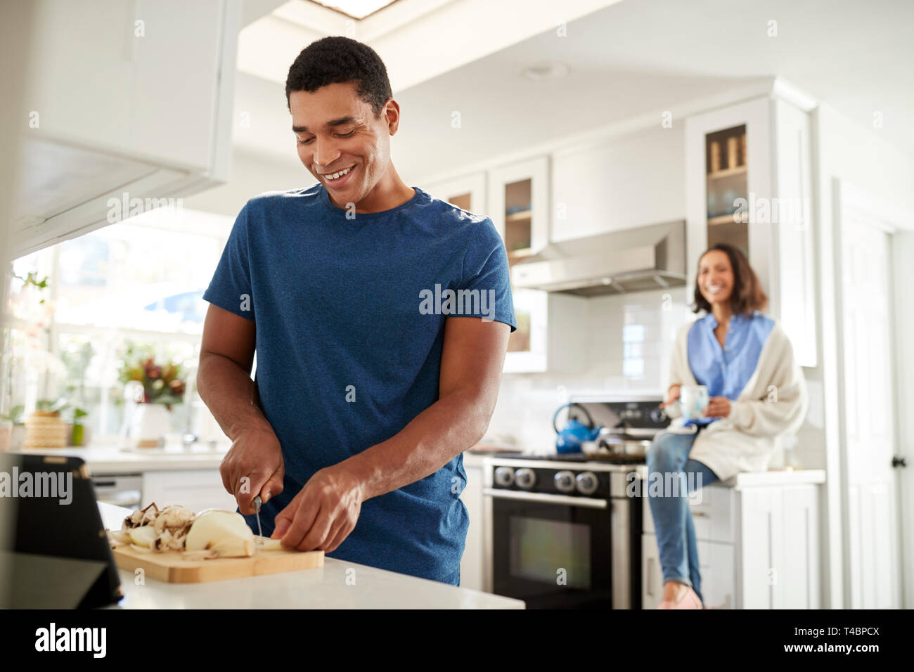 Jeune adulte homme debout dans la cuisine la préparation des aliments, son partenaire assis sur le plan de travail de cuisine derrière lui, l'accent sur le premier plan Banque D'Images
