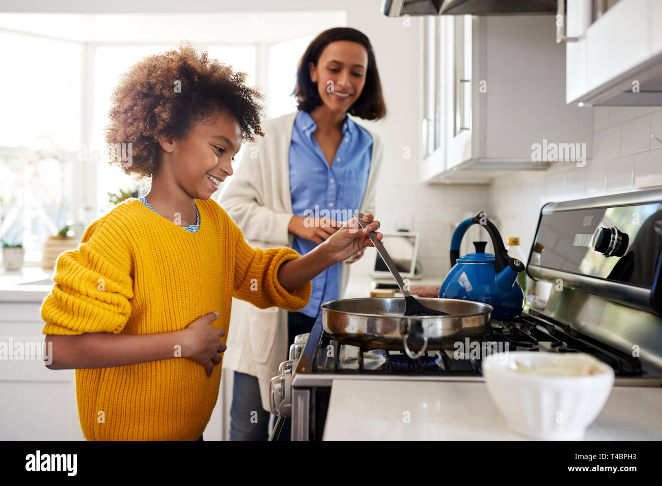 Fille préadolescente debout à plaque de cuisson dans la cuisine à l'aide de spatule et poêle, préparer la nourriture avec sa mère, side view Banque D'Images