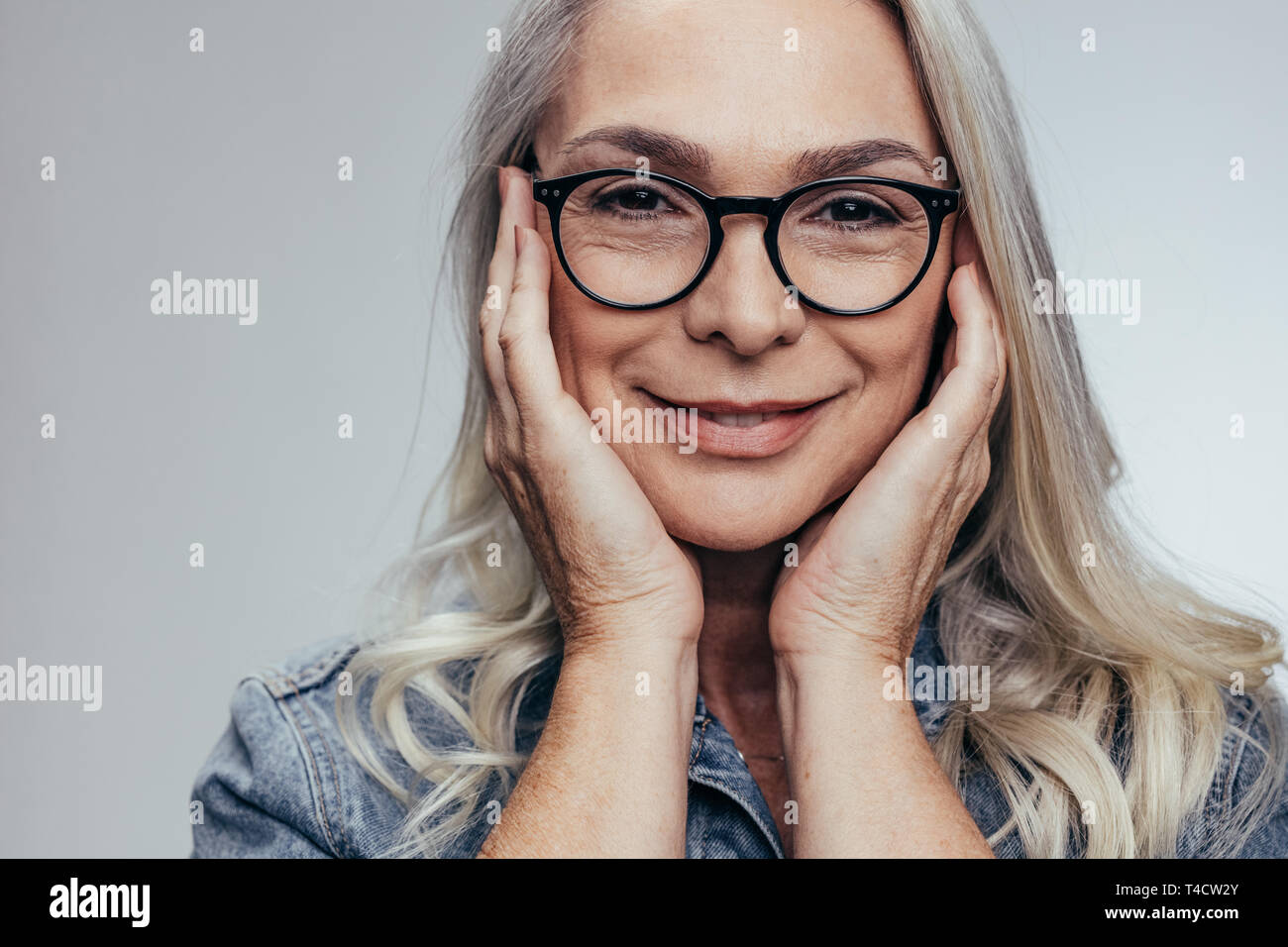 Senior woman avec les mains sur son visage contre l'arrière-plan gris. Mature positive woman in casual chemise en jean. Banque D'Images