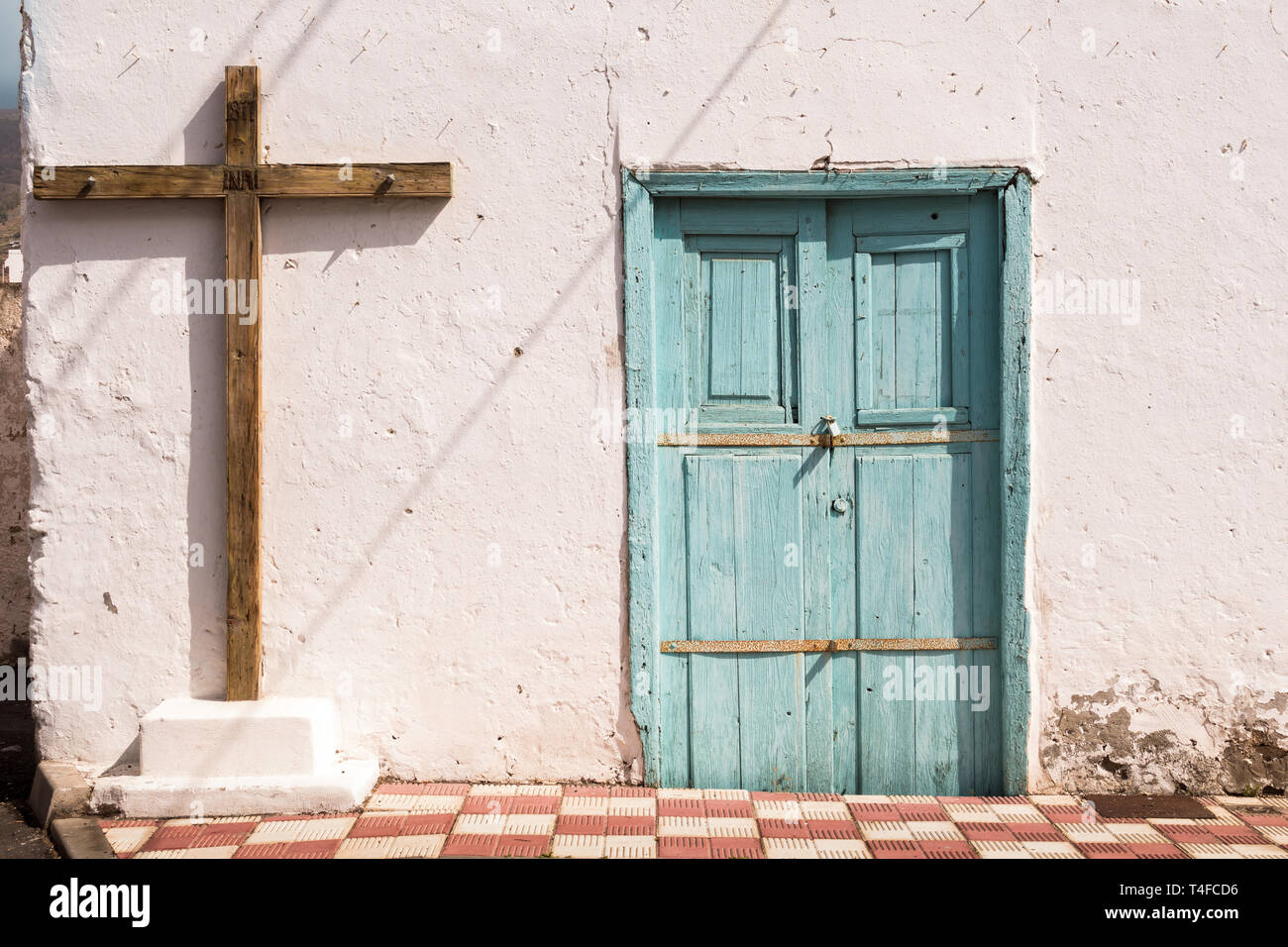 Éclairé lumineux mur d'une maison. Vieille porte en bois bleu et une croix. Les ombres des câbles en diagonale sur le mur. Chaussée d'échecs rouge et blanc. Arico Banque D'Images