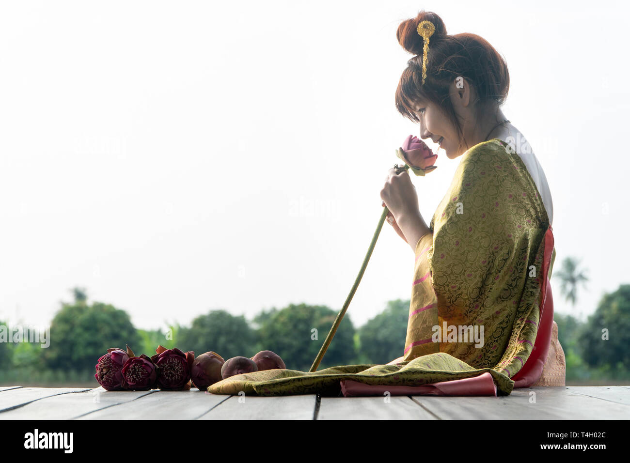 Belle Asie femmes portant robe traditionnelle thaïlandaise et assis sur un plancher en bois. Ses mains se tenant fleur de lotus et l'odeur de fleur rose. Banque D'Images