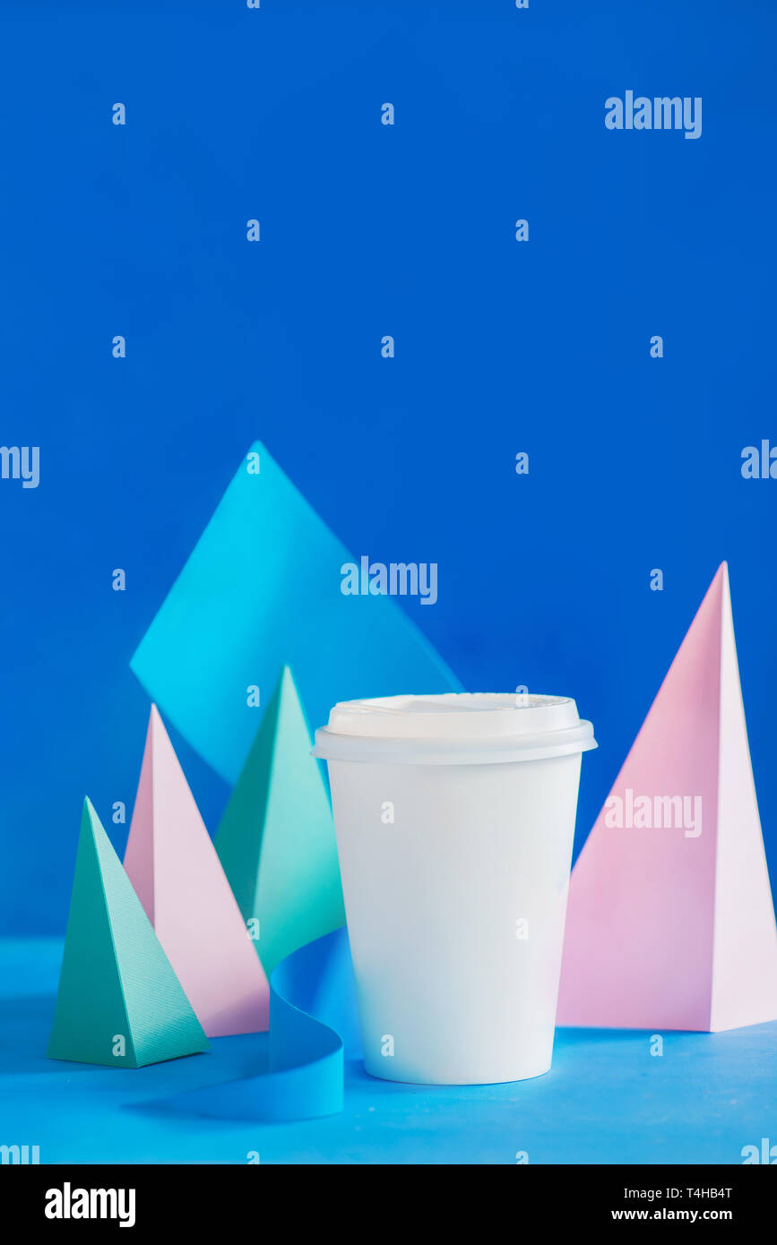 Le papier blanc tasse pour le café sur un fond abstrait avec sculpture papier moderne. Pyramides de papier et des courbes design dans des couleurs pastel. Banque D'Images