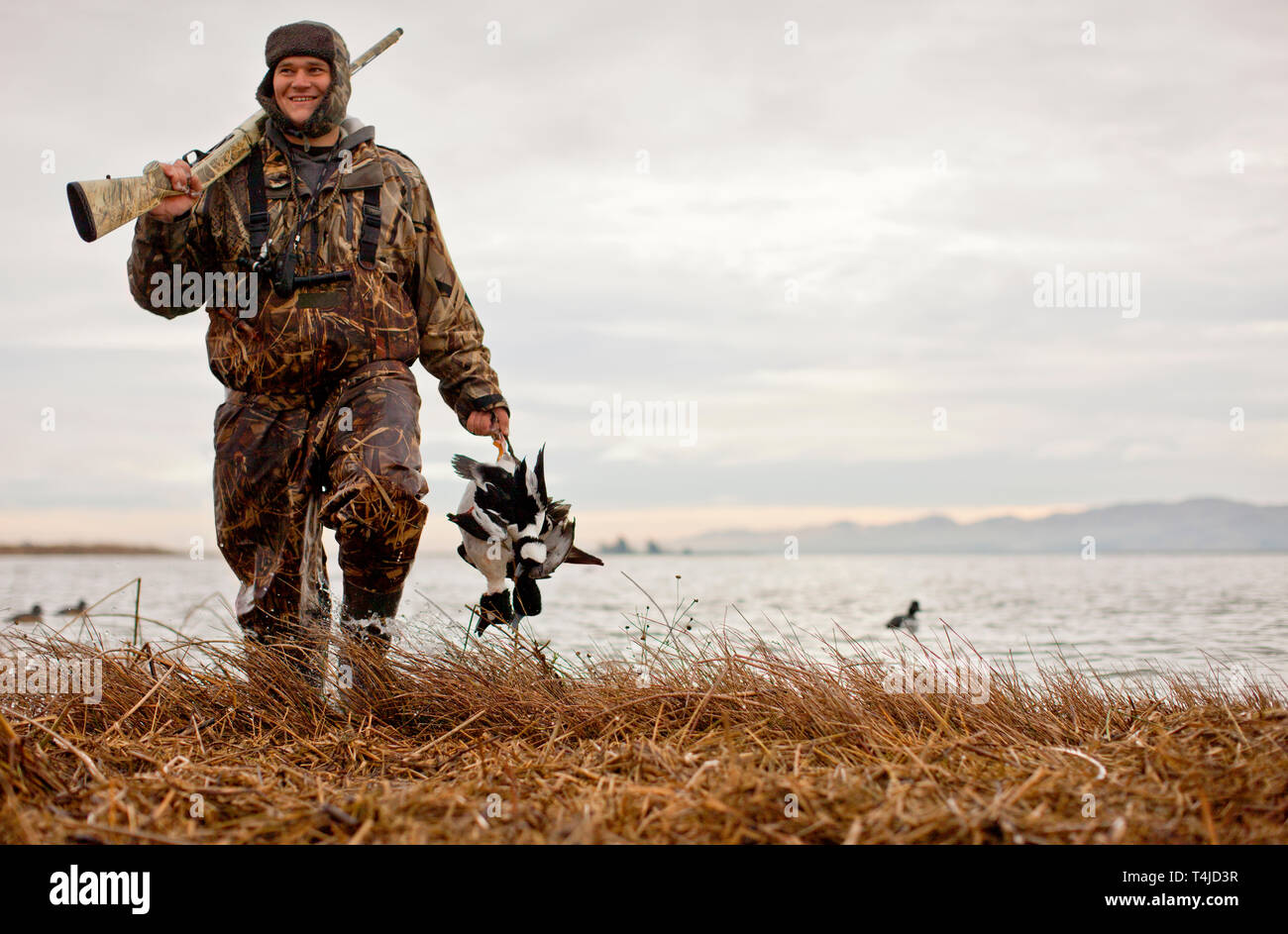 Satisfait chasseur de canard portant des vêtements de camouflage avec deux canards morts et fusil pendant qu'il marche le long du bord d'un lac. Banque D'Images