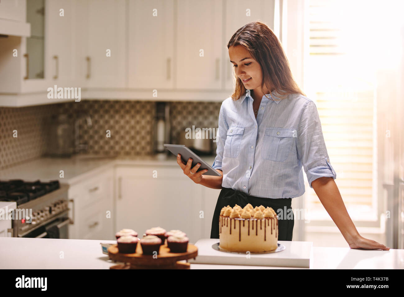 Femme debout dans la cuisine à l'aide de tablette numérique avec articles de pâtisserie sur le comptoir de la cuisine. Femme chef looking at her tablet pc dans la cuisine. Banque D'Images