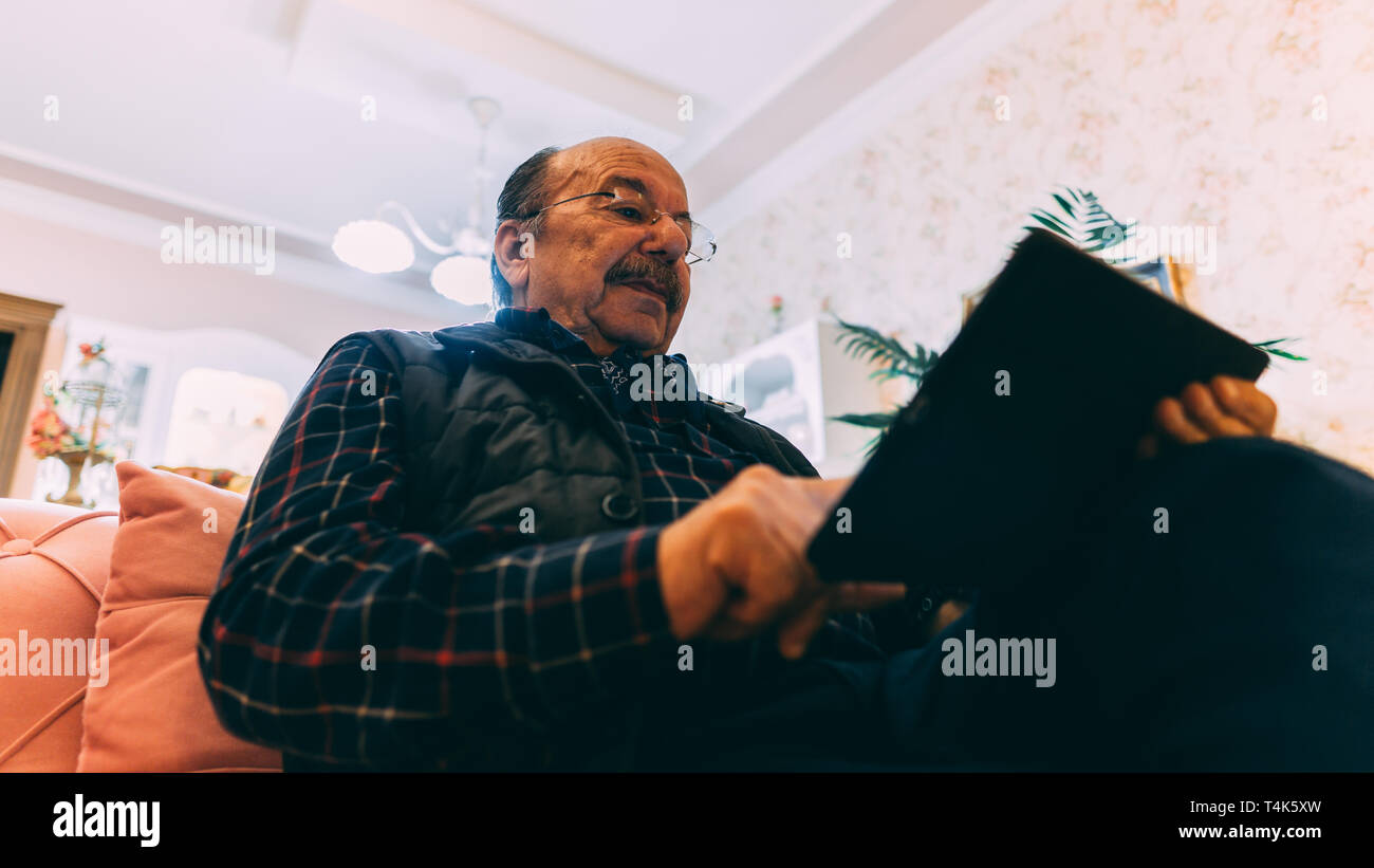 Senior man with tablet sitting on sofa at home.old man en utilisant sa tablette. Low angle view. Banque D'Images Senior man with tablet sitting on sofa at home.old man en utilisant sa tablette. Low angle view. Banque D'Images