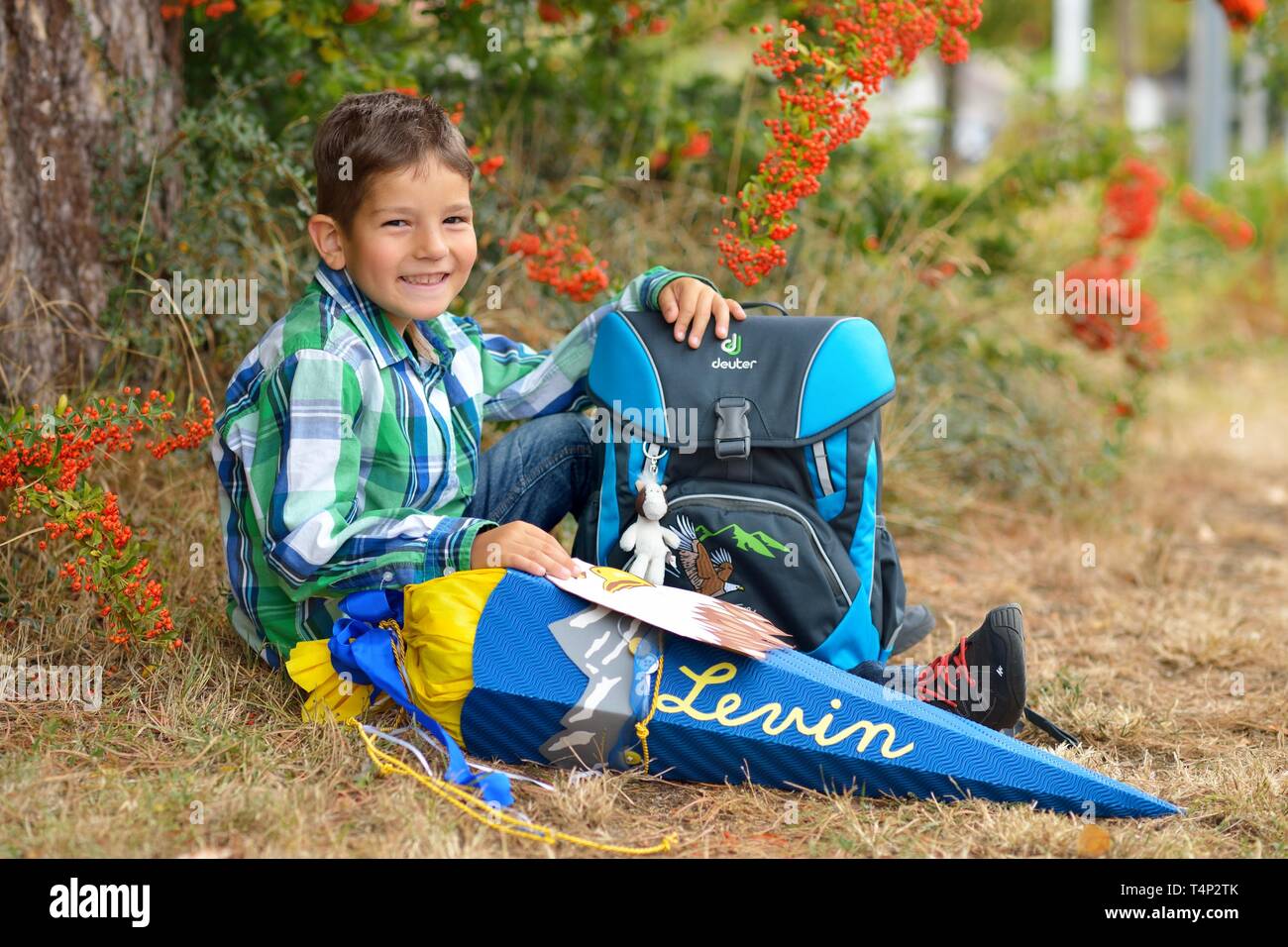 Garçon, 6 ans, premier jour d'école, s'assied et sourit avec sac d'école et de l'école à l'arbre conique, Allemagne Banque D'Images