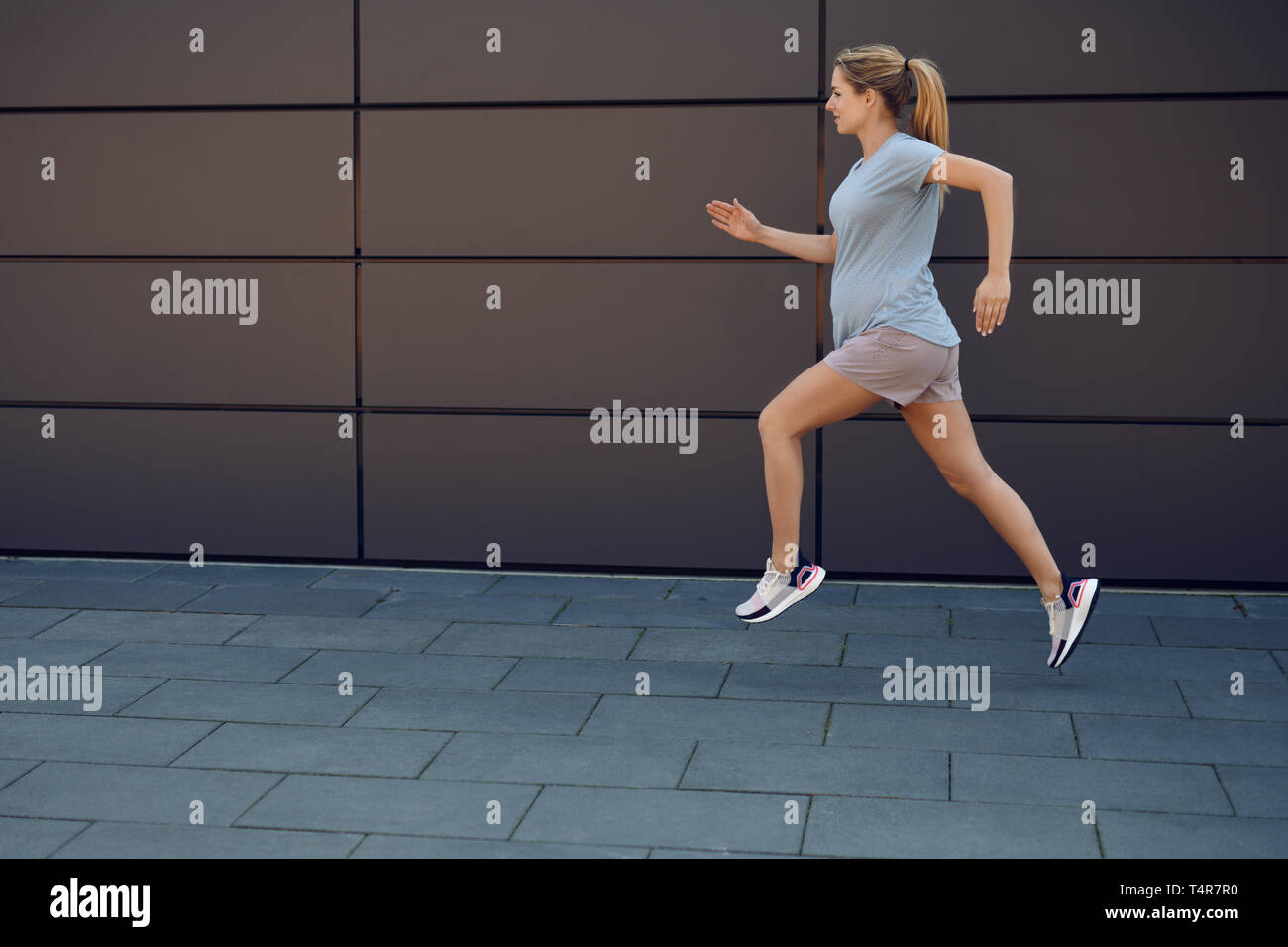 Jeune femme enceinte keeping fit courir le long d'un trottoir pavé en face d'un mur urbain dans un concept de vie sain Banque D'Images