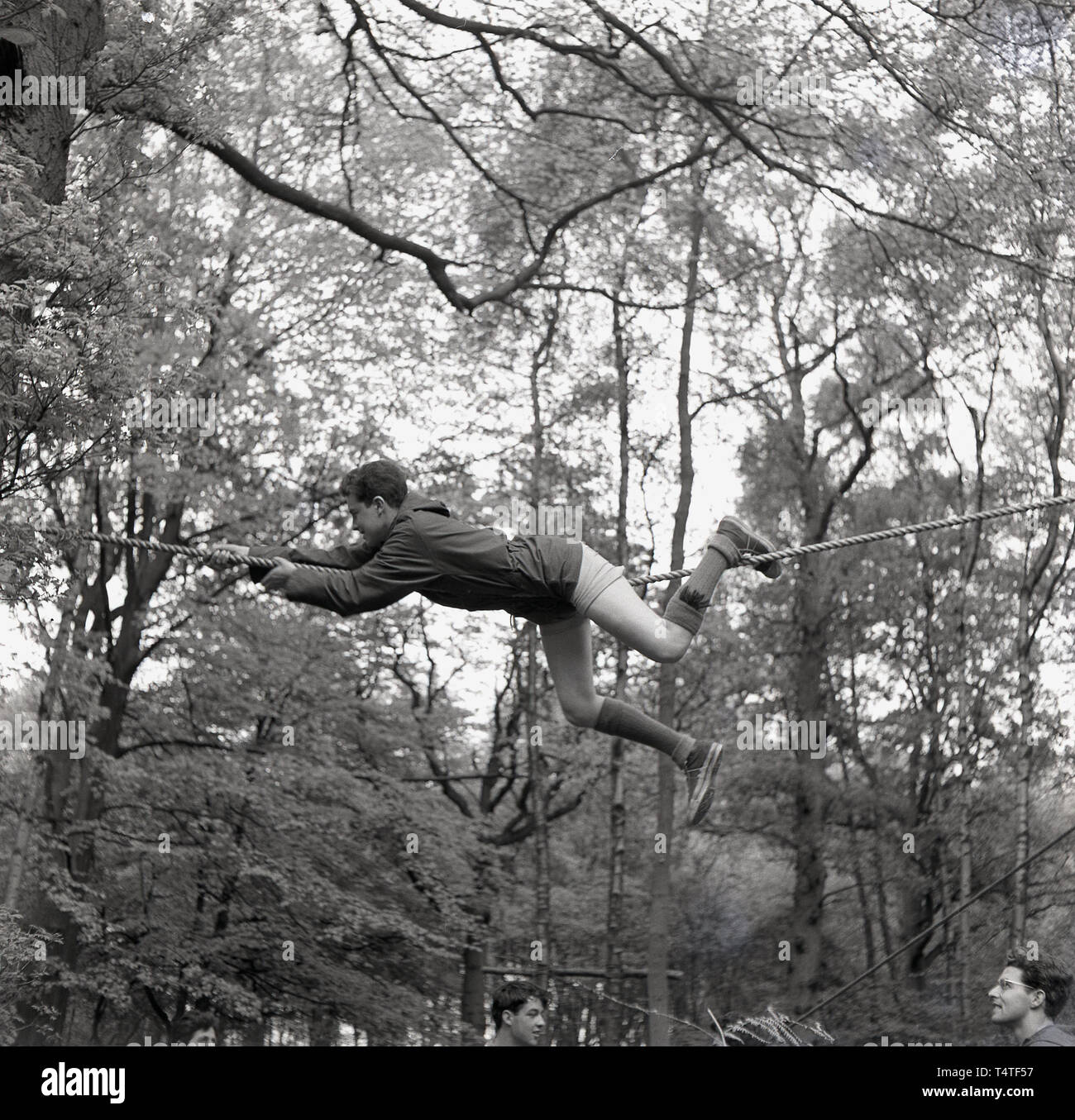 Années 1960, historique, un jeune scout aventure couchée sur une corde à l'aide de ses mains et pieds de manœuvre lui-même à travers une clairière entre deux arbres, England, UK. Les jeunes dans les scouts prennent part à de nombreuses activités de plein air qui contribuent à développer leur caractère et à la vie quotidienne. Banque D'Images