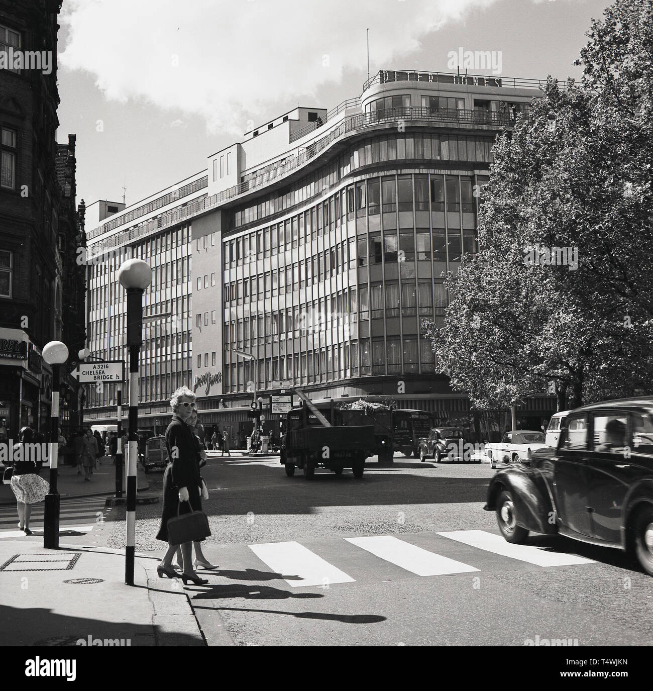 Années 1960, historiques, deux dames en traversant un passage à niveau à la station Sloane Square, Chelsea, Londres, avec la grande façade de verre, grand magasin Peter Jones, fondée en 1877, sur la rue Kings Road, vu dans la distance. Banque D'Images