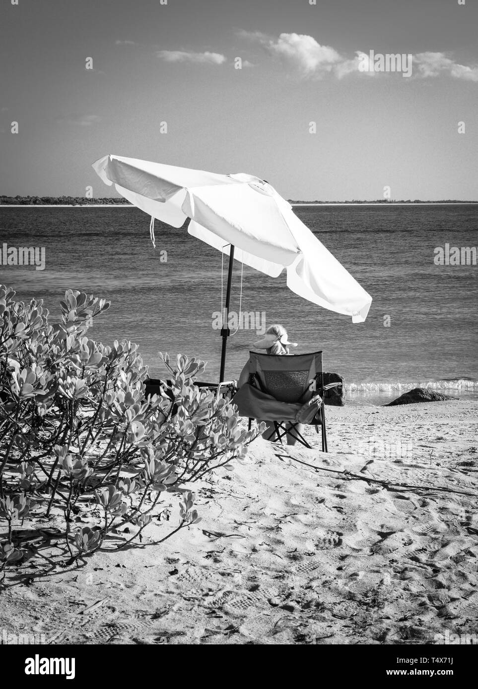 Une vue arrière d'une blonde woman relaxing dans une chaise de plage dans un cadre donnant sur le bord de l'eau bleue de son perchoir en noir et blanc Banque D'Images