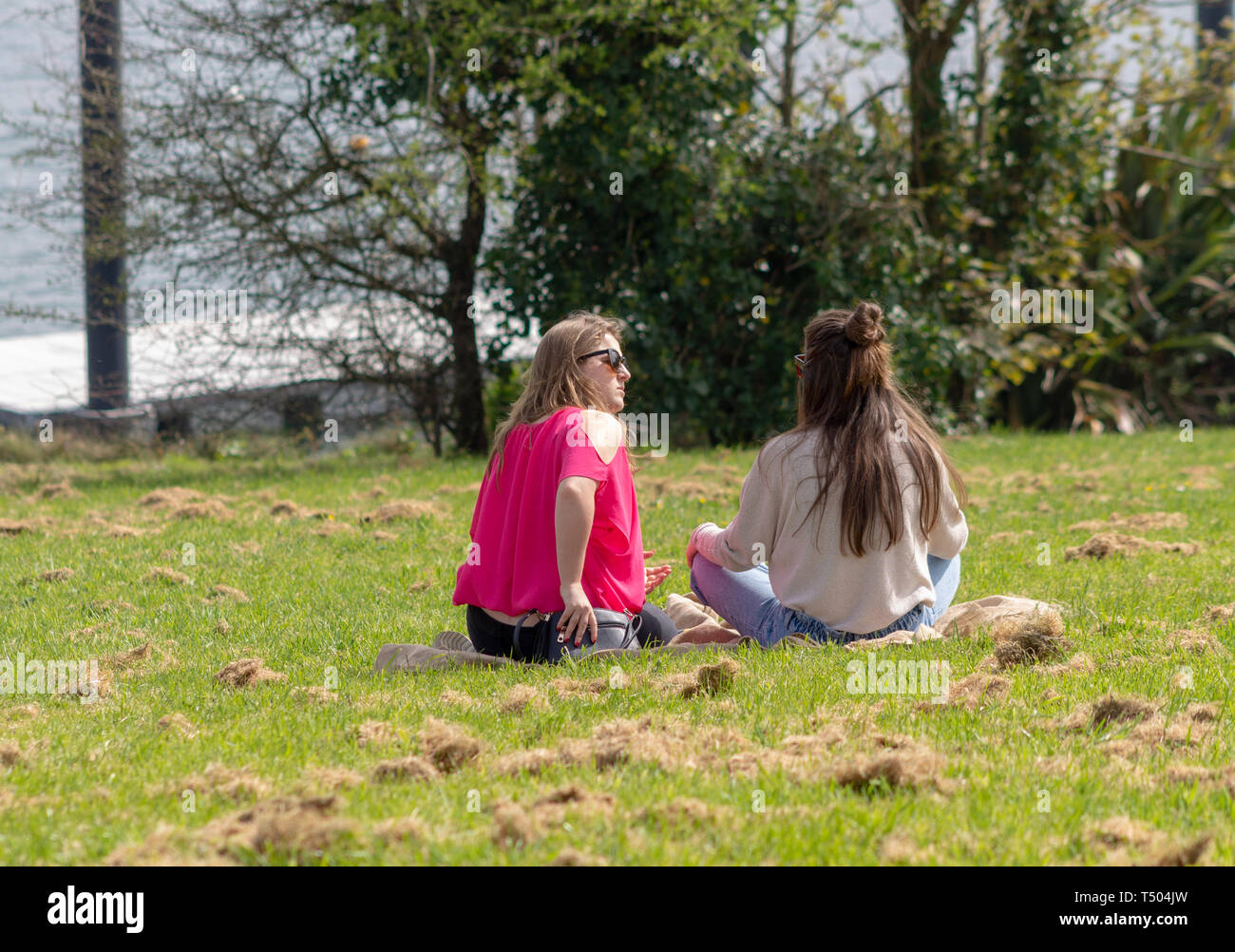 2 jeunes femmes filles s'assit sur l'herbe dans un parc, profitant du soleil. Banque D'Images