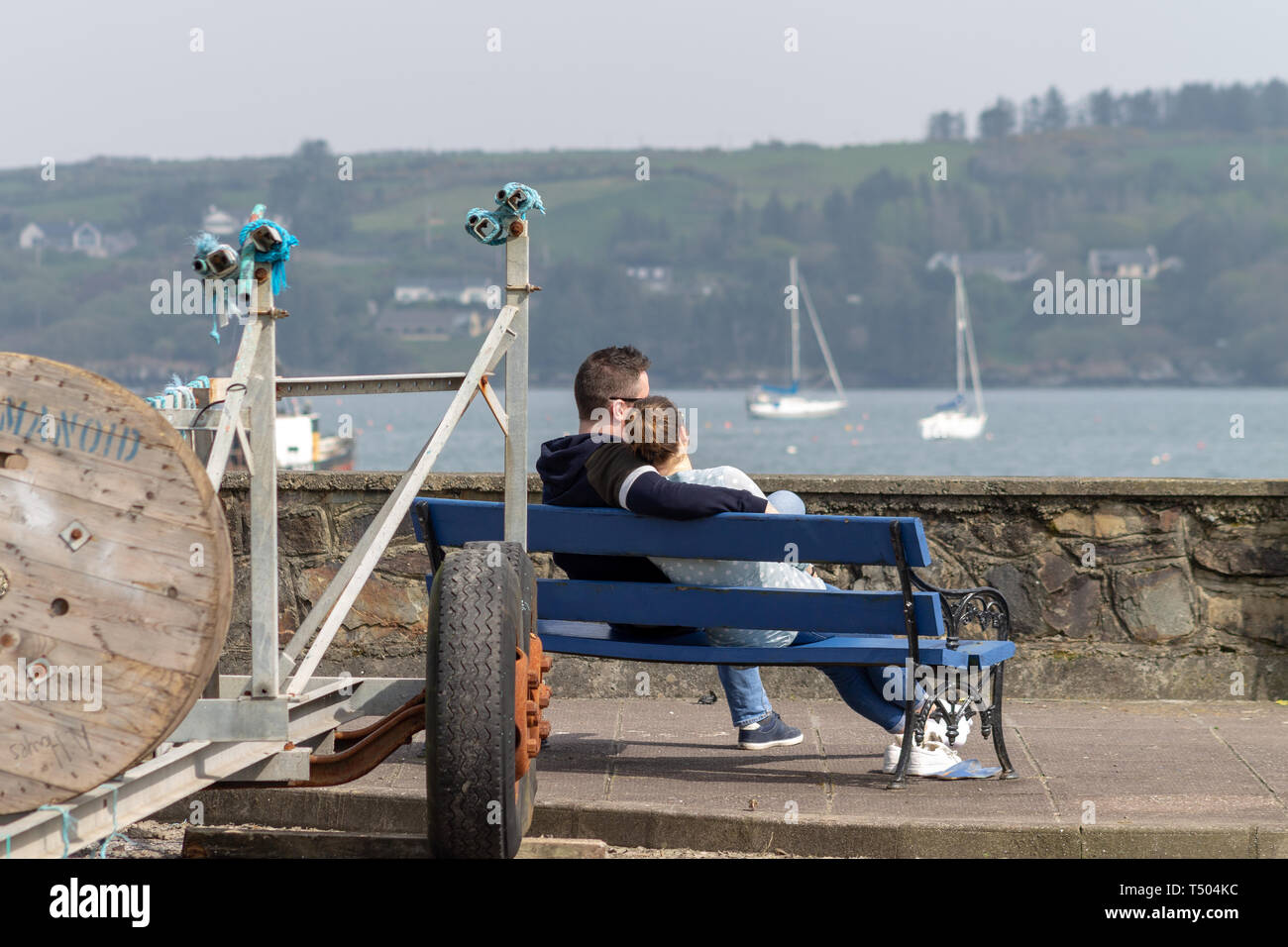 Jeune couple assis sur un banc de parc par la mer dans les bras appréciant chaque compagnie d'autres personnes dans le soleil Banque D'Images