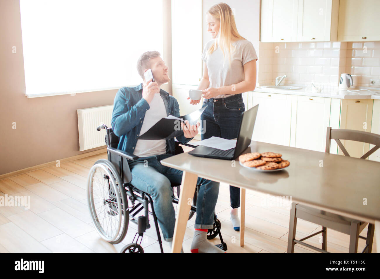 Jeune homme gai avec l'inclusion s'asseoir à table et parler au téléphone. Il regarde la femme et sourire. Holding dossier ouvert et l'étude. Je coupe modèle tenir Banque D'Images