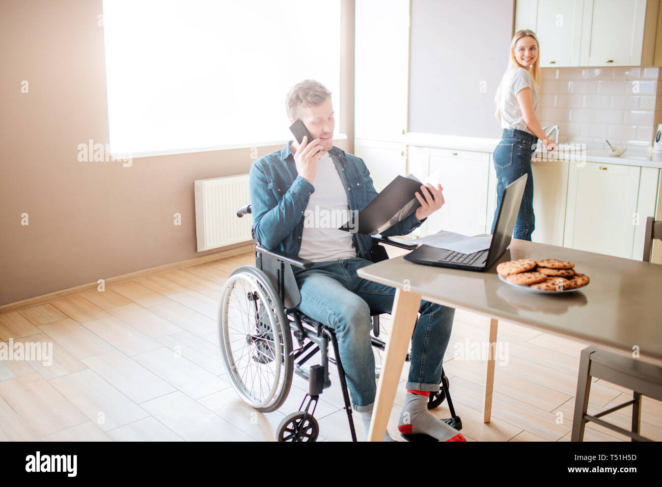 Jeune étudiant en fauteuil roulant occupé sur l'étude et de prendre sur le téléphone. Guy ayant des besoins spéciaux et d'invalidité. Holding dossier ouvert. Jeune femme stand à stov Banque D'Images