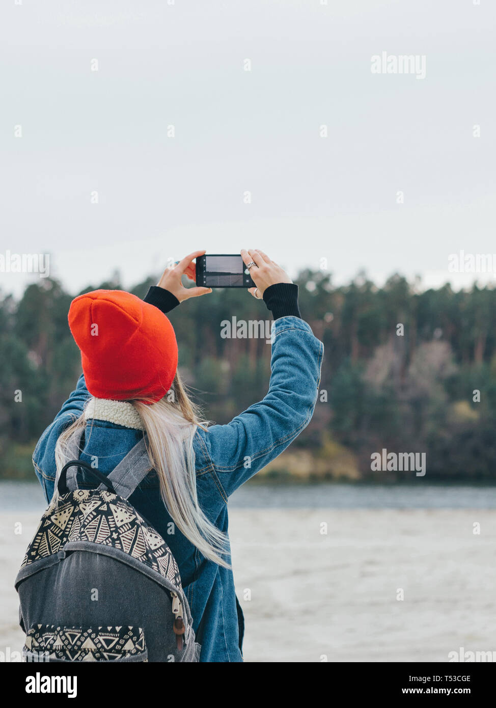 Promenade le long de la rivière, une jeune fille dans un bonnet rouge avec  un sac à dos est de prendre des photos de la rivière sur un smartphone  Photo Stock -