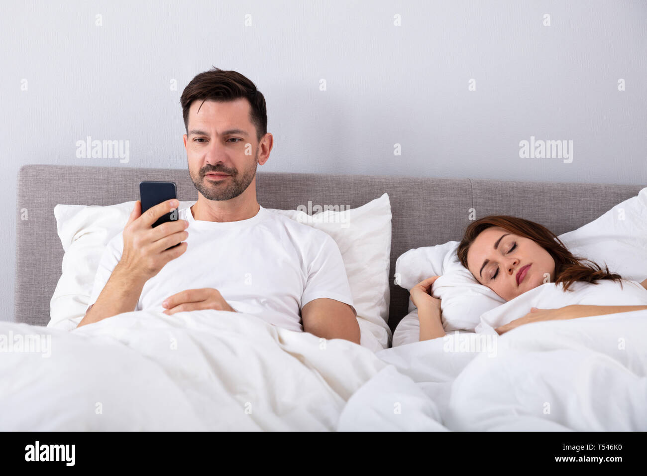 Smiling Man Using Smartphone pendant que sa femme dort sur le lit dans la chambre Banque D'Images