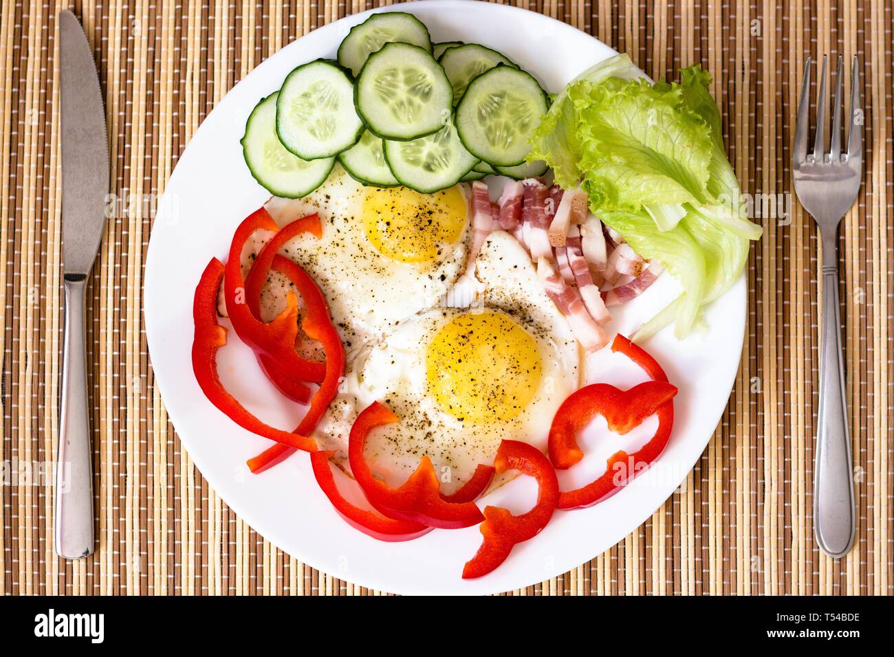 Poulet frit délicieux et frais oeufs frais avec du poivre rouge, du bacon, de la laitue et le concombre. Le plat est sur une plaque blanche avec un couteau et fourchette. Banque D'Images