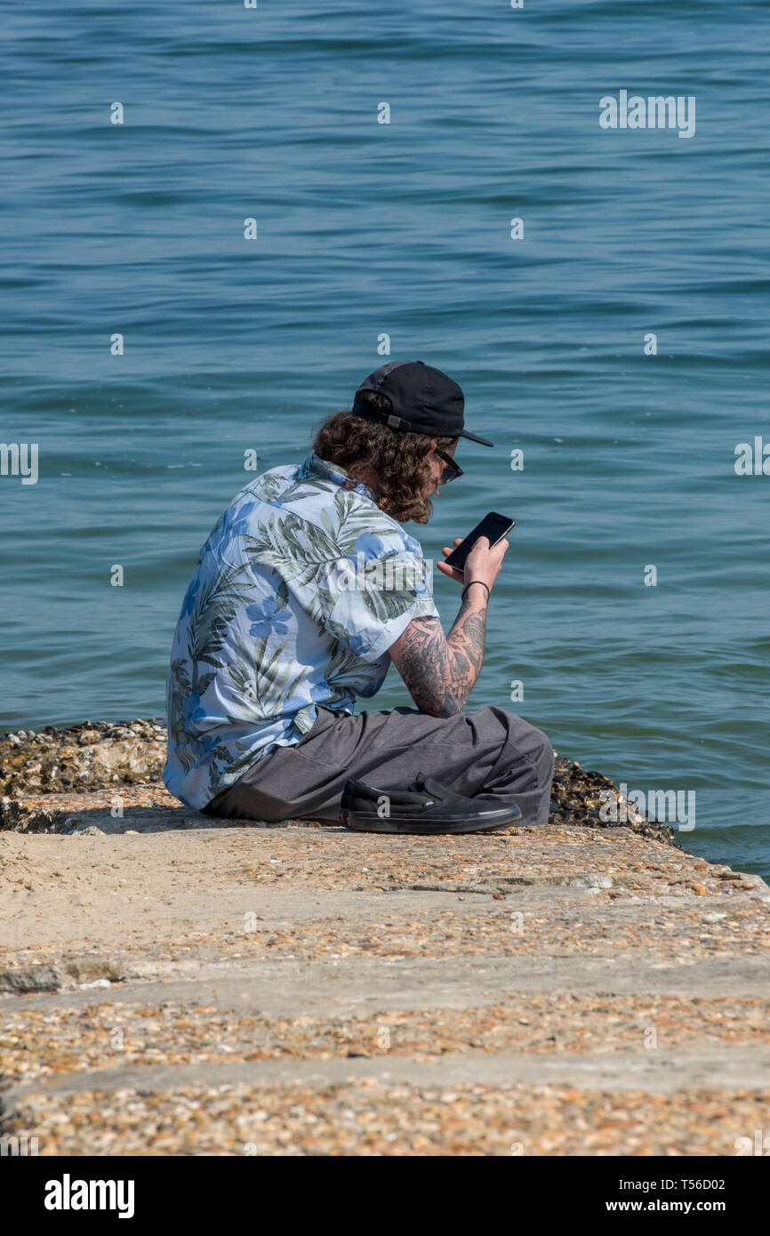 Jeune homme ou une jeune adolescente aux cheveux longs assis au bord de la mer bord de l'eau à l'aide d'un smartphone téléphone mobile portant casquette de baseball. Banque D'Images