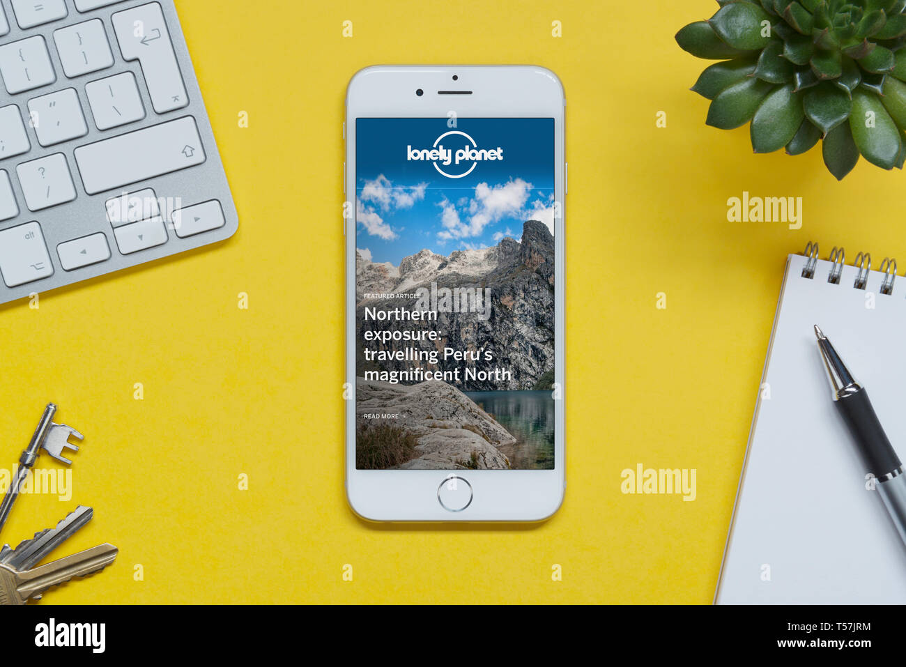 Un iPhone montrant le site Lonely Planet repose sur un fond jaune table avec un clavier, les touches, le bloc-notes et des plantes (usage éditorial uniquement). Banque D'Images