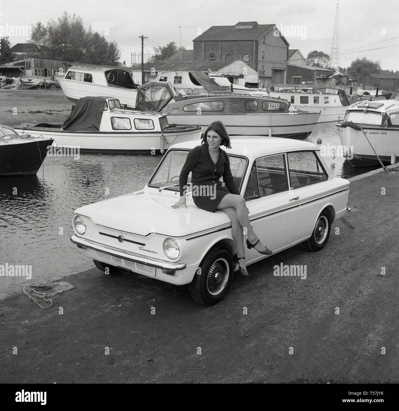 1967, historique, à l'extérieur dans un chantier naval, une jeune femme portant un cardigan en laine et une jupe assise sur le coffre d'une voiture Sunbeam Sport, la version sportive de la célèbre petite voiture, la Hillman Imp. La Hillman Imp était une petite voiture fabriquée par Rootes Group et son successeur Chrysler Europe de 1963 à 1976. C'était le concurrent dans la catégorie des petites voitures de la British Leyland Mini. Banque D'Images