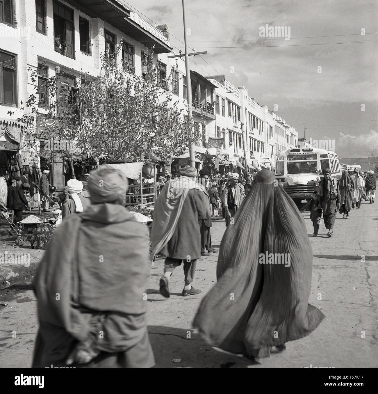 Années 1950, Kaboul, Afghanistan, les afghans en costume national dans une large rue poussiéreuse passant les étals du marché et les bus dans le centre-ville. Banque D'Images