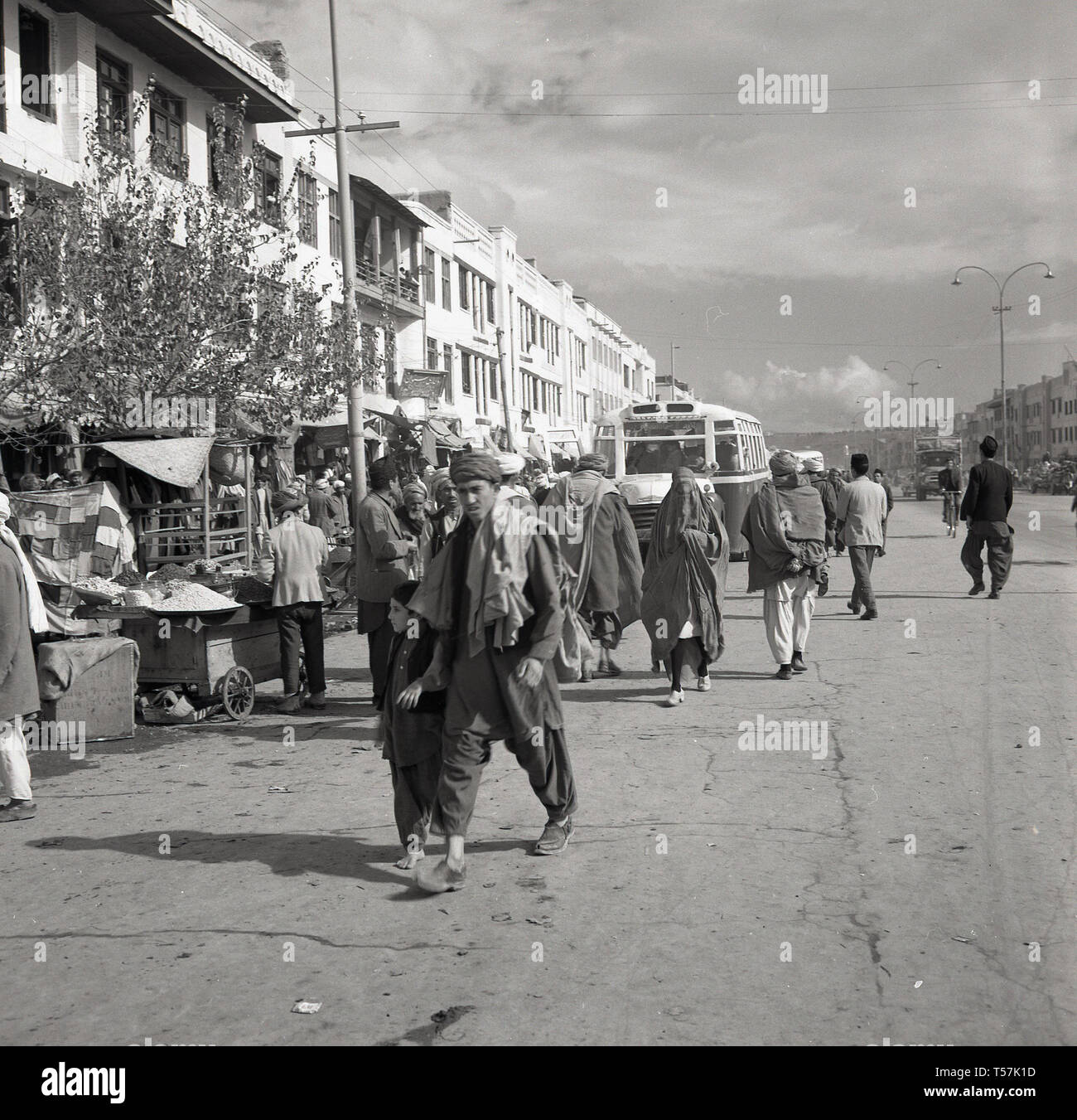 Années 1950, Kaboul, Afghanistan, les afghans en costume national dans une large rue poussiéreuse passant les étals du marché dans le centre-ville. Banque D'Images