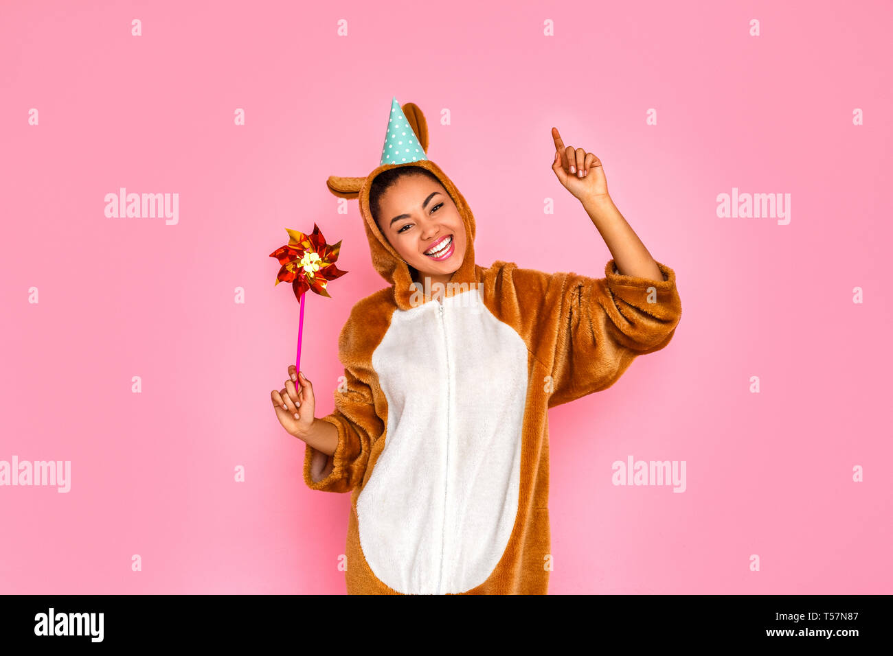 Jeune femme en bunny kigurumi wearing party hat standing isolé sur fond rose holding pinwheel posant à l'appareil photo au space pinting up smiling che Banque D'Images
