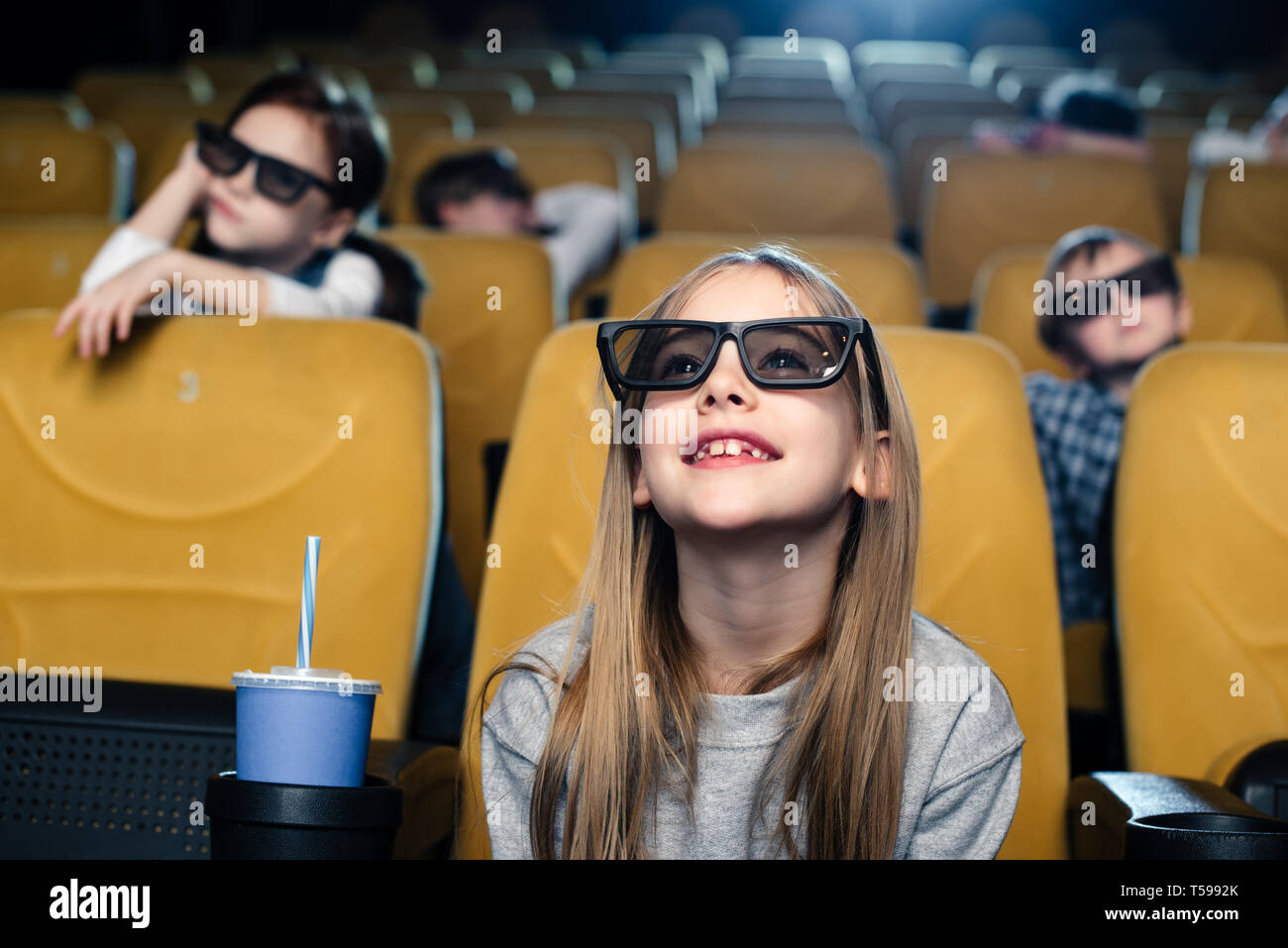 Smiling blonde enfant dans les lunettes 3d assis près de tasse de papier et regarder des films dans le cinéma Banque D'Images