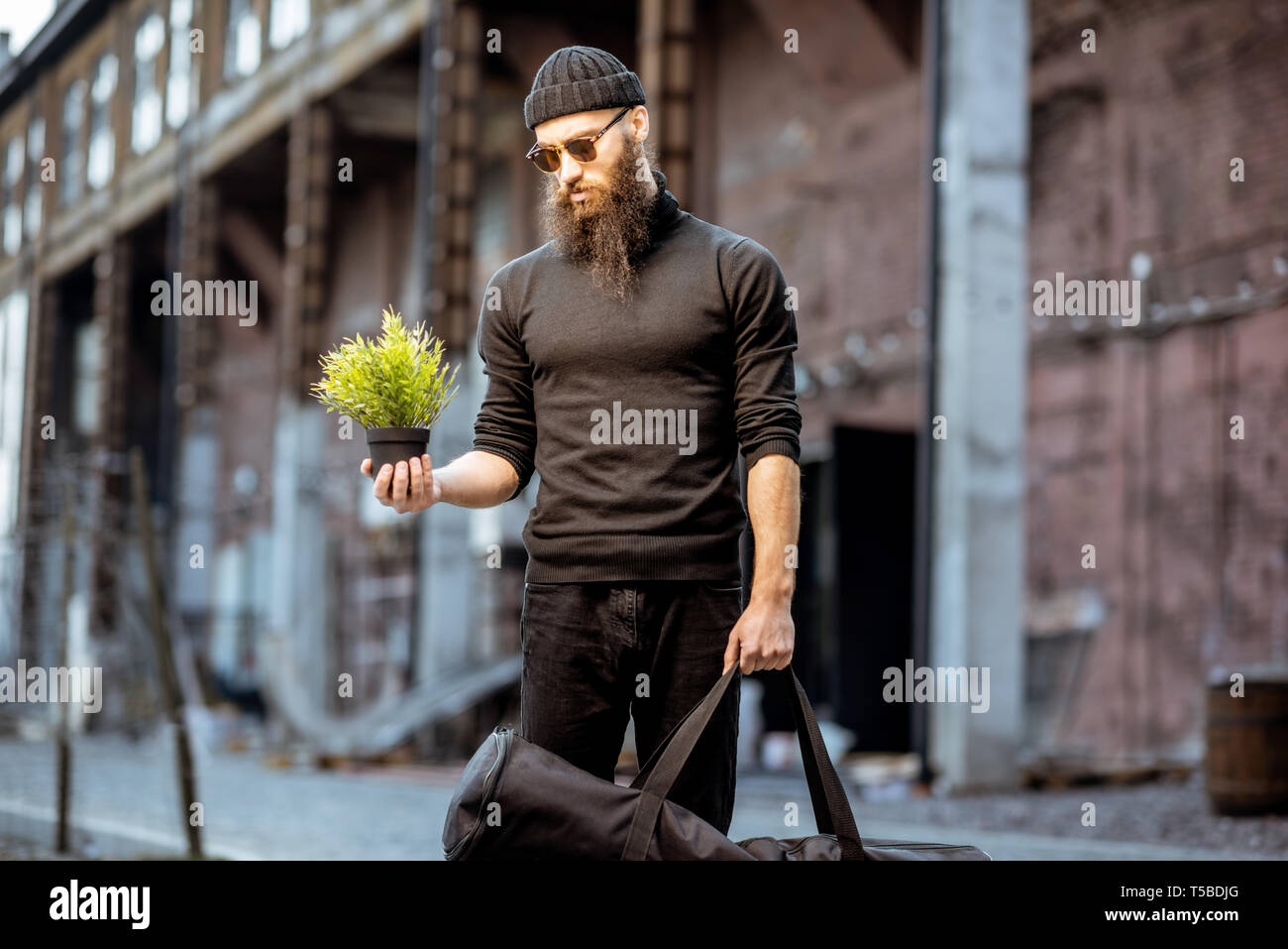 Portrait d'un homme barbu sérieux comme un tueur vêtu de noir des vêtements serrés holding pot de fleurs et sac avec arme à l'extérieur Banque D'Images