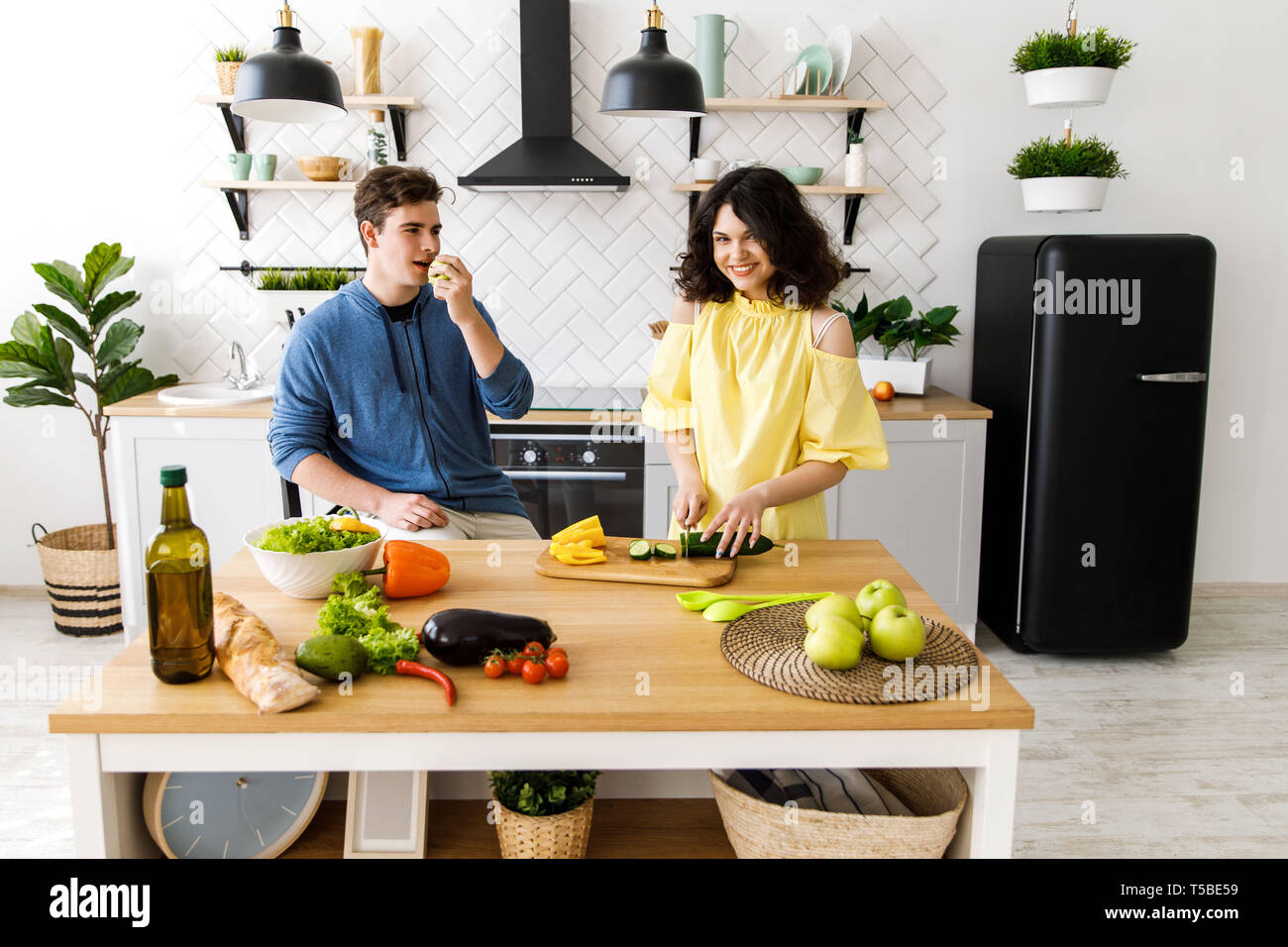 Un couple - une fille et un garçon qui passent du temps ensemble pour la cuisine. Le mec mange de la pomme verte, la fille coupe légumes pour une salade Banque D'Images