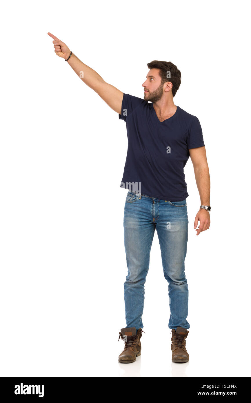 Bel homme sérieux en jeans, bottes et t-shirt bleu est debout, pointant vers le haut et en montrant quelque chose. Studio pleine longueur shot isolé sur blanc. Banque D'Images