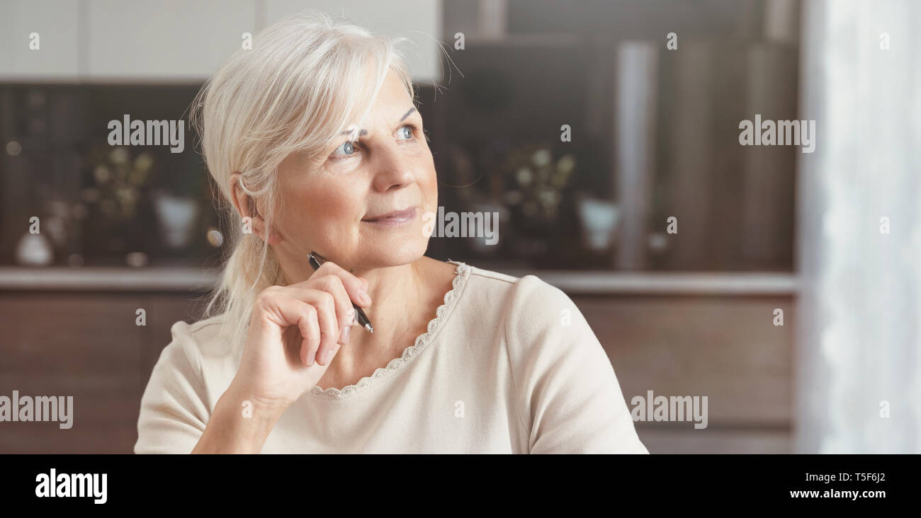 Cheerful senior woman portrait. Pensionné woman holding a pen, est assis à la maison et regarde la fenêtre. Concept d'une heureuse retraite Banque D'Images