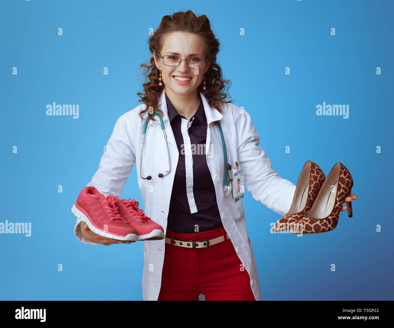 Femme médecin élégant souriant en bue shirt, pantalon rouge et blanc robe médical fitness montrant les espadrilles et hautes chaussures de talon isolé sur bleu. Banque D'Images