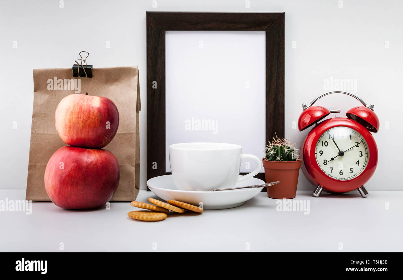 Foncé maquette, cadre photo réveil rouge, sac de papier, deux pommes, les craquelins et une tasse de café blanc avec une soucoupe sur un fond clair. Banque D'Images