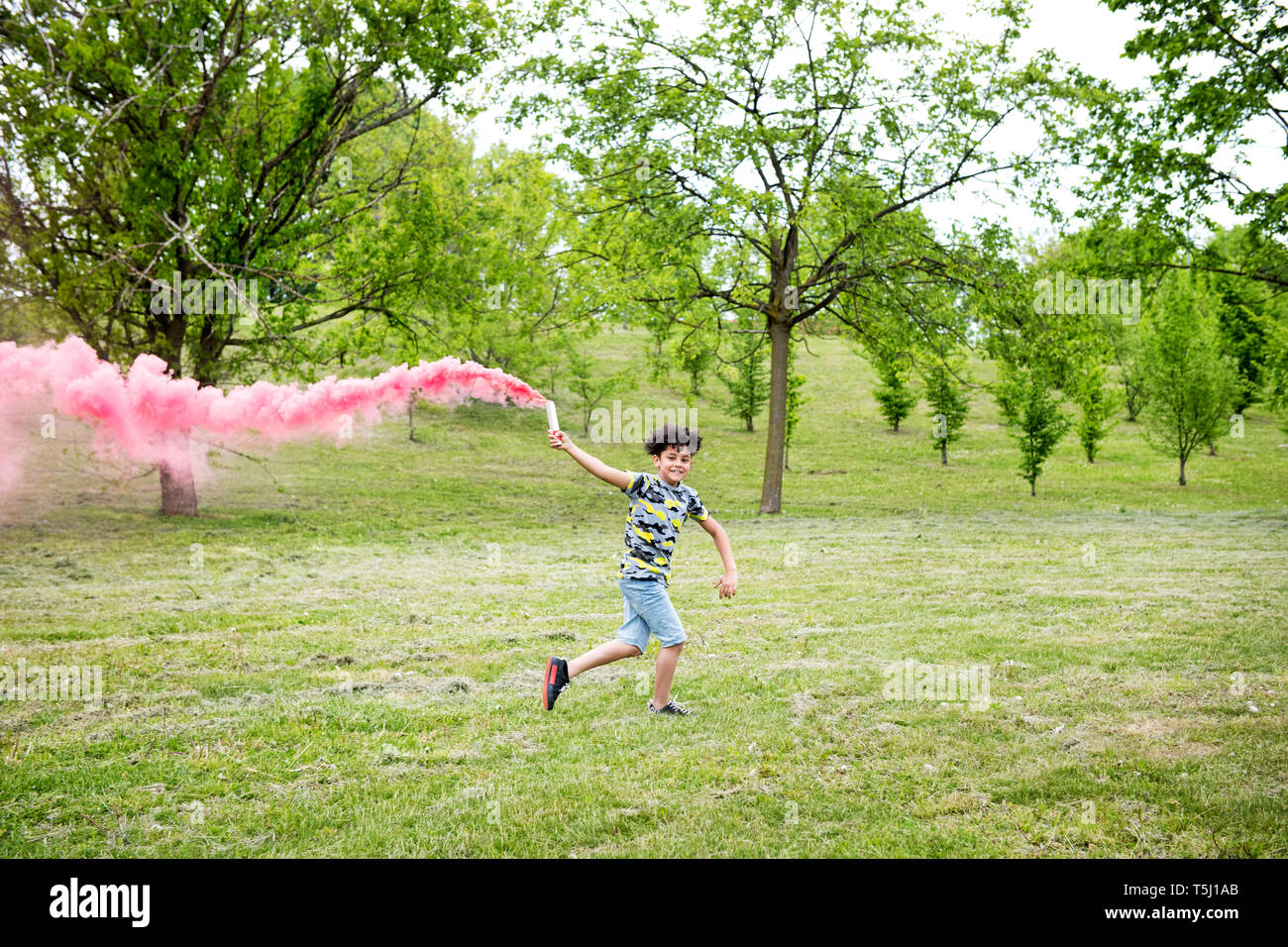 Jeune garçon courir avec un flare rose coloré de fuite de fumée rose derrière lui dans l'herbe dans un parc aux arbres de ressort Banque D'Images