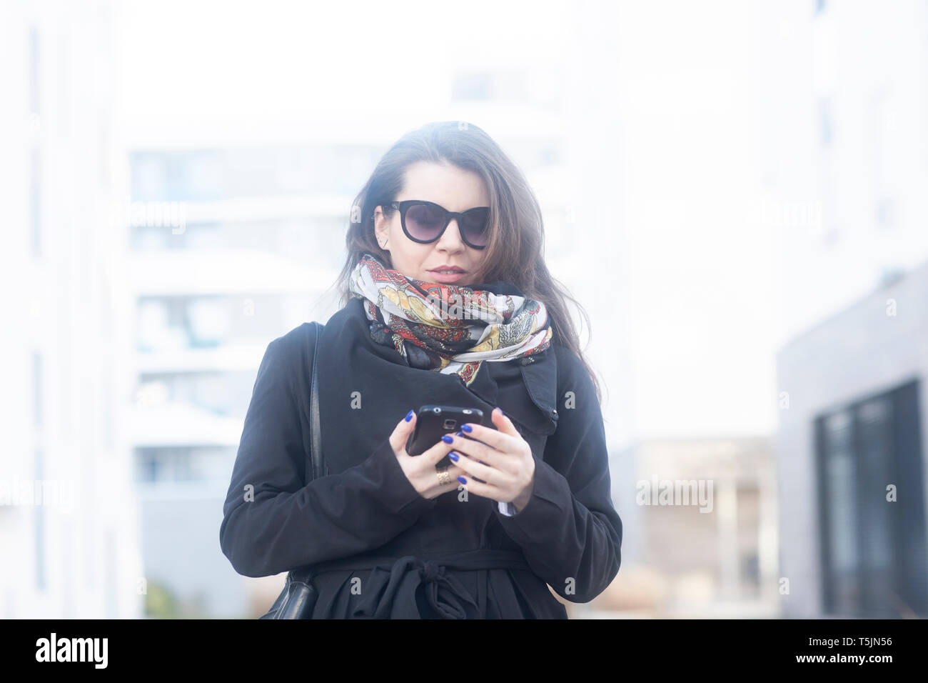 Portrait de jeune femme portant manteau, écharpe et lunettes looking at cell phone Banque D'Images