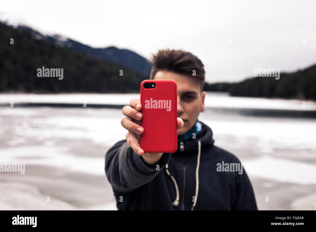 Jeune homme tenant téléphone cellulaire rouge à un lac en hiver Banque D'Images